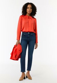 Czerwona satynowa bluzka z marszczoną dekoltem, zestawiona z dopasowanymi ciemnoniebieskimi dżinsami i beżowymi szpilkami, trzymająca pasującą czerwoną marynarkę.