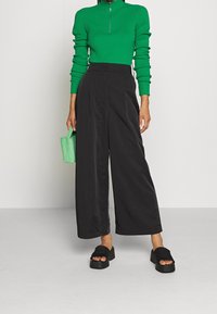 Pantalon noir à jambes larges avec plis sur le devant, associé à un pull vert côtelé à col zippé. Sandales à plateforme noires et petit sac à main vert complètent la tenue.