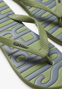 Grønne og marineblå flip-flops med en tekstureret sål. Stropperne er grønne, med et fiskebensmønster og mærkenavnet "Superdry" præget.