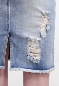 Jupe en denim bleu clair avec ourlet effiloché, fente avant et plusieurs zones déchirées aux bords bruts. Design décontracté, longueur genou.