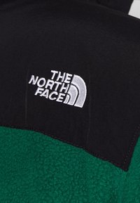 Fleece bunda se zeleným tělem a černým horním panelem, s vyšívaným bílým logem "The North Face" na hrudi.