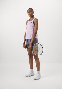 Lilla sporttank top, millel on teemantmuster ja logo, koos tumedate lühikeste pükstega. Mudel hoiab tennisereketit, jalas on valged spordijalatsid ja sokid.