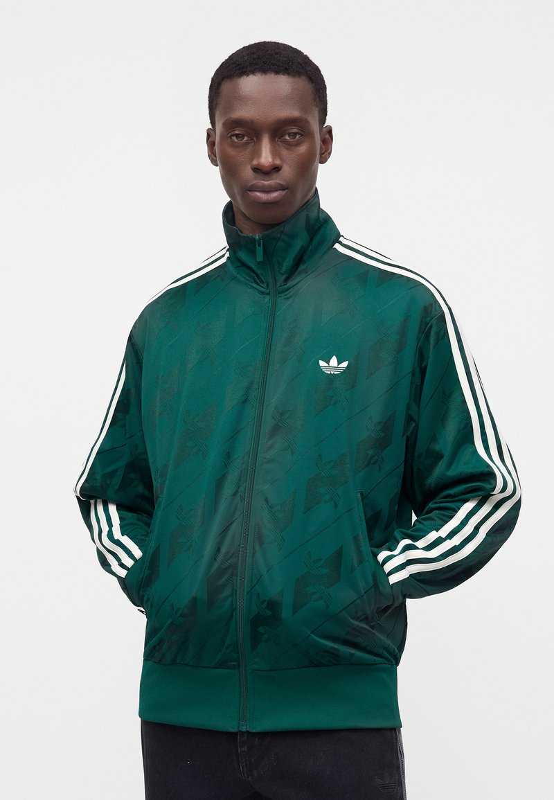 Hombre con chaqueta deportiva Adidas verde oscuro con rayas blancas en las mangas y manos en los bolsillos, mirando hacia adelante sobre un fondo liso.