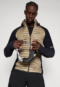 Casaco acolchoado, em tons de bege e preto, com capuz e fecho éclair, apresentando um padrão acolchoado. A pessoa segura uma alça com um padrão em preto e branco.