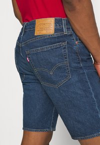 Tmavě modré džínové šortky s rovnýcm střihem, které mají koženou nášivku v pase a žluté prošití na zadních kapsách.