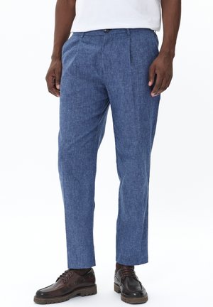 Pantalons bleus sur mesure en tissu léger, avec un devant plissé, des jambes droites et une fermeture à bouton. Portés avec des chaussures brunes.