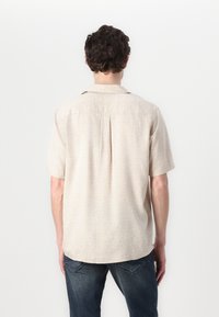 Camisa de manga corta beige con un diseño estampado, detalle en el cuello y corte relajado. Combinada con jeans azules. Se muestra vista trasera.