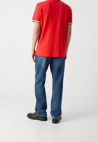 Mężczyzna ubrany w czerwoną koszulę polo z krótkim rękawem, niebieskie dżinsy i czarne buty, stojący na tle białej ściany, odwrócony plecami.