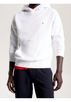 Mann trägt einen weißen Hoodie mit Bauchtasche, kleinem Logo auf der Brust, rotem Shirt darunter und dunkler Hose vor einfachem Hintergrund.