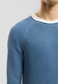 Blauer gestrickter Pullover mit strukturiertem Muster, rundem Ausschnitt und Raglanärmeln, über einem weißen Unterhemd getragen.
