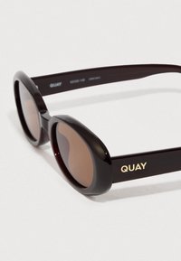 Fekete, túlságosan nagy méretű napszemüveg barna lencsékkel, ívelt kerettel és arany "QUAY" logóval az oldalon. Simább műanyag anyag.