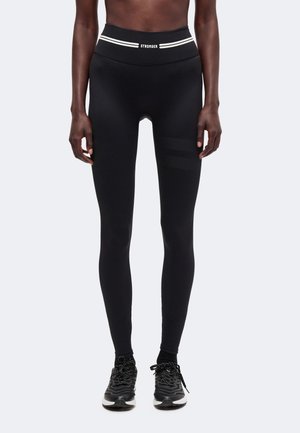 Person iført sorte højtaljede leggings med teksten "STRONGER" på linningen og sorte sportssko mod en ensfarvet baggrund.