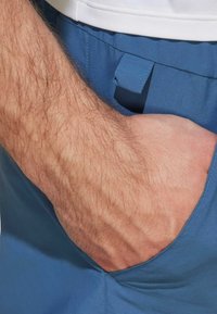 Blå shorts med en slät textur, med en sidficka och en tygögl. Närbild av en hand som vilar i fickan.