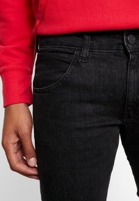 Černé džíny z denimu s úzkým střihem, které mají přední kapsy, zapínání na knoflíky a hladkou texturu. Pas je mírně zvýšený.