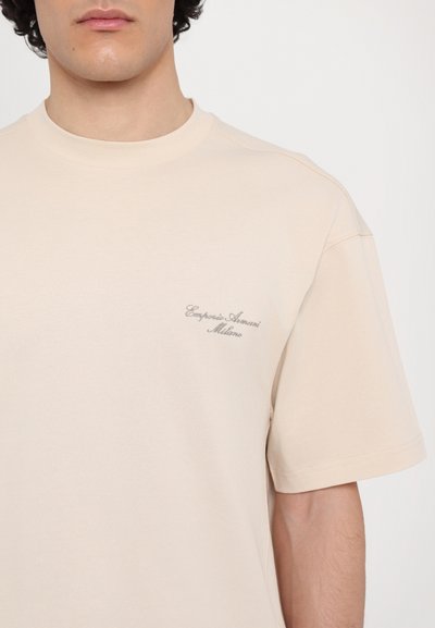 Béžové bavlněné tričko s kulatým výstřihem a krátkými rukávy. Na levé hrudi se nachází jemně vyšívané logo. Hladká textura, minimalistický design.