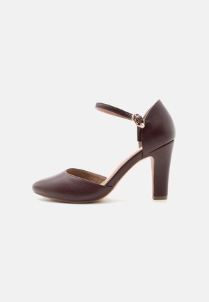 Scarpa con tacco alto in pelle marrone scuro con punta chiusa e appuntita, cinturino alla caviglia con fibbia dorata e dettaglio con perla, tacco a blocco.