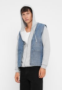 Vest din denim albastru cu două buzunare frontale, suprapus peste un hanorac gri cu glugă și fermoar pe față, asortat cu pantaloni negri.