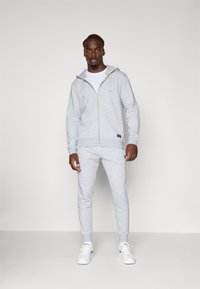 Lys grå zip-up hættetrøje med frontlommer og matchende grå sweatpants. Glat overflade, ribbede manchetter og hvide sneakers fuldender outfittet.