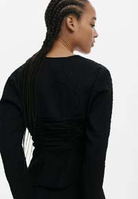 Top nero texturizzato con maniche lunghe, caratterizzato da un design arricciato sul retro. Il tessuto ha una superficie decorata con sottili variazioni di texture.