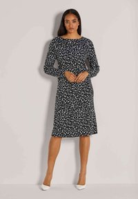 Frau steht gegen eine beige Wand und trägt ein langärmliges, knieumspielendes Kleid mit schwarz-weißen Leopardenmuster und weiße spitze Absätze.