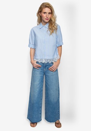 Blonde Frau trägt eine hellblaue, gestreifte Kurzarmbluse mit Spitzenbesatz, weite blaue Jeans und braune Sandalen, steht mit den Händen in den Taschen.