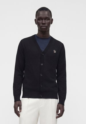 Mand iført sort knap-op cardigan med lille broderet zebra-logo, marineblå skjorte og hvide bukser, stående mod en ensfarvet baggrund.