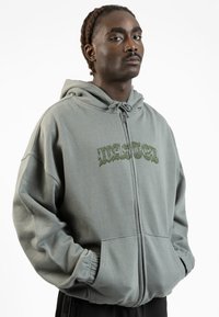 Felpa con zip verde in cotone, dal taglio loose, con cappuccio regolabile e tasche frontali. Presenta la scritta "WASTED" stampata in verde scuro.