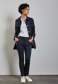 Impermeable de color azul marino con grandes botones plateados sobre una camisa blanca, combinado con pantalones slim de color azul marino y zapatillas blancas. Textura suave, diseño clásico.