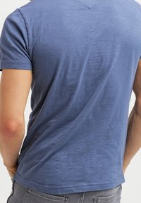 Hombre usando una camiseta azul ajustada de manga corta y jeans grises, visto desde atrás sobre un fondo simple.