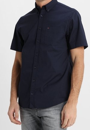 Mann trägt ein marineblaues, kurzärmeliges Hemd mit Knöpfen und kleiner Brusttasche sowie hellgraue Jeans.