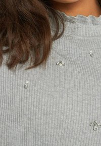 Pull gris à côtes avec un col à volants, orné de petites perles et de cristaux éparpillés sur le tissu.