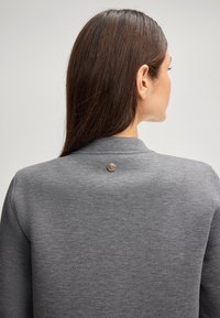 Maglione grigio con una texture liscia, caratterizzato da un dettaglio con bottone metallico rotondo nella parte posteriore del collo. Maniche lunghe e design minimale.