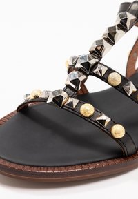 Sandalia de cuero negro con tachuelas metálicas y acentos dorados texturizados. Suela plana con un patrón trenzado a lo largo del borde.