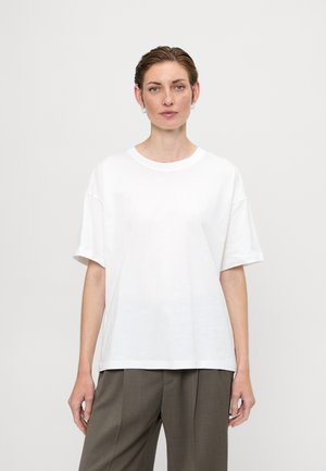 Weißes Baumwoll-T-Shirt mit entspanntem Schnitt, Rundhalsausschnitt und kurzen Ärmeln, kombiniert mit grauen, maßgeschneiderten Hosen. Glatte Textur, schlichtes Design.