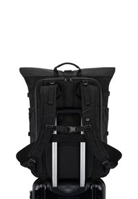 Fekete hátizsák, párnázott pántokkal, rögzítve egy fekete kerekes bőrönd fogantyújára, hátulról látható fehér háttér előtt.