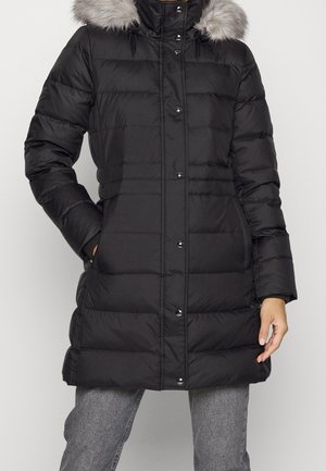 Manteau d'hiver noir matelassé avec boutons-pression, poches latérales et capuche doublée en fausse fourrure grise, porté avec un jean gris.