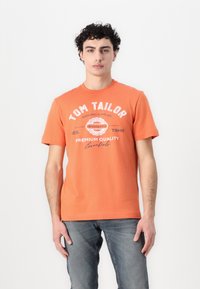 Oranžs kokvilnas t-krekls ar īsām piedurknēm, kuram priek�špusē ir balti drukāti teksti un apļa logotips. Valkāts ar ziliem džinsiem.