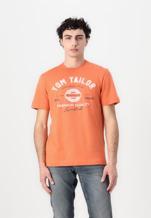 Camiseta de algodón naranja de manga corta, con texto impreso en blanco y un logotipo circular en el pecho. Combina con pantalones vaqueros azules.