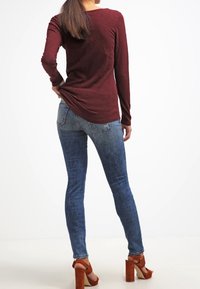 Kvinna med långt brunt hår som bär en vinröd långärmad topp, blå skinny jeans och bruna högklackade sandaler, stående vänd bort. 