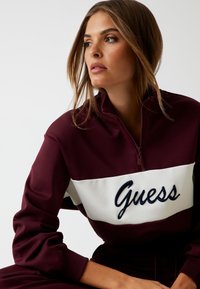 Felpa zip in borgogna con una banda orizzontale bianca e il logo "Guess" ricamato in blu navy. Tessuto morbido, vestibilità comoda e polsini a coste.