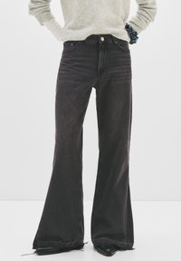 Personne portant un jean noir taille haute et large avec des ourlets effilochés, un pull gris clair et des chaussures noires à bouts pointus sur un fond uni.