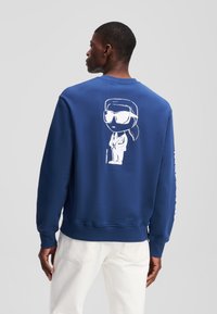 Felpa navy con grafica di un personaggio dei cartoni animati sul retro, polsini e orlo a costine. Realizzata in morbido tessuto, presenta una vestibilità comoda.