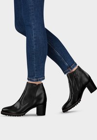 Des jambes portant un jean slim bleu foncé et des bottines noires en cuir à talons blocs avec des fermetures éclairs sur le côté, le tout sur un fond clair.