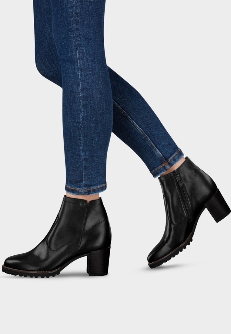 Des jambes portant un jean slim bleu foncé et des bottines noires en cuir à talons blocs avec des fermetures éclairs sur le côté, le tout sur un fond clair.