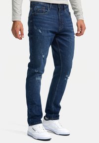 Vaqueros de mezclilla azul oscuro con detalles desgastados, corte slim, diseño de cinco bolsillos, combinados con zapatillas blancas y una camiseta de manga larga gris claro.