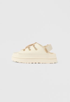 Sandalia beige con plataforma plana, puntera abierta y cordones trenzados. Fabricada con un material liso, suela texturizada y correa ajustable.