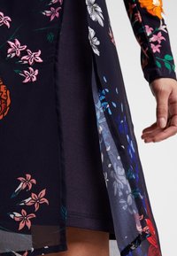 Robe maxi florale avec des fleurs brodées roses et oranges sur un fond bleu marine, avec un revêtement en tissu transparent et une fente sur le côté. Texture lisse.