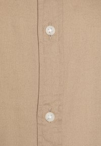 Beige button-up shirt with a textured fabric, featuring two white buttons and a side seam, highlighting a simple and clean design.