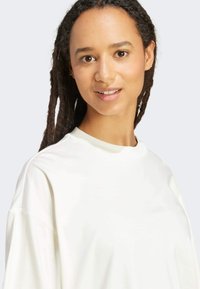 White, lightweight sweatshirt with crew neckline, dropped shoulders, and smooth texture. Model has long, dark, curly hair and a neutral background.