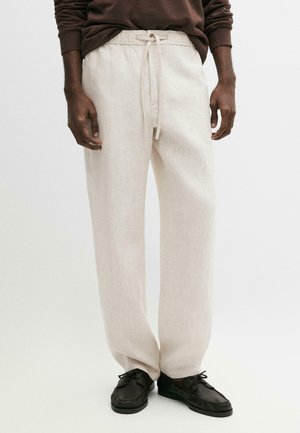 Mann trägt beige Leinenhose mit Kordelzug, dunkelbraunes Langarmshirt und schwarze Schnürschuhe vor weißem Hintergrund.